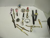 Wide view showing all 16 wristwatches arranged on white surface, variety of bands and face shapes.