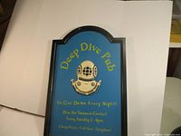 Front view of the blue vintage pub sign with rounded black frame, yellow lettering, and raised diver helmet.
