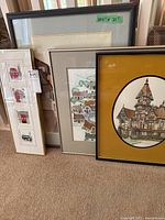 Framed artworks grouped on the floor showing size and style variations, including a large one with a label 20.5 x 27 and smaller vertical frames with British themes.