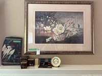 Framed Asian themed artwork above shelf with assorted decorative items including tins, boxes, a clock and ceramic hanging