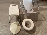 Photo showing metal frame shower chair with plastic backrest and padded armrests alongside beige plastic toilet seat cover and beige over toilet raised seat on concrete floor.