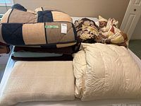 Photo showing folded bedding items including a blue and beige comforter, floral comforter, and light colored duvet on top of a bed.