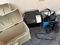 Wide view showing large beige plastic storage box, black file folder box, blue file folder container with compartments, two cameras in separate cases, and binoculars on carpeted floor.