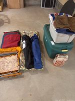 Photo showing folded cushions, air mattress bags, tote bags, large plastic storage bins, and bag of BBQ charcoal briquettes arranged on basement floor.