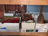 Boxes of Christmas tree balls in silver, gold, and red colors, along with faux poinsettia flowers, stacked on a basement shelf.