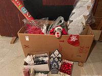Cardboard box containing assorted Christmas decorations such as wrapping paper, plush ladybug, artificial red berries, white angel decoration, and red stocking decoration.
