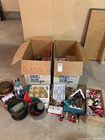 Two full boxes of assorted Christmas ornaments and decorations, including glass baubles in red and green, decorative figurines, and small holiday decor items arranged on the floor.