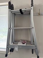Collapsible aluminum ladder folded and hung against the wall showing overall structure and fold strap.