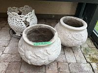 Photo of two concrete planters side by side on a brick floor with a white decorative ceramic pot holder behind.