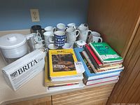 Overview of stacked cookbooks, Brita filter jug with box and new filters, and multiple ceramic cups arranged on a kitchen counter