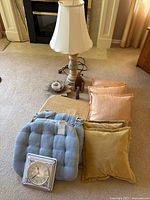 Photo showing assorted cushions including blue tufted chair cushions, yellow and peach decorative pillows, beige chair cushion, lamp with white shade, flower candle holder, clock and figurines on floor.