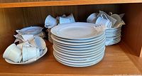 Set of 12 white dinner plates stacked with white porcelain cups around them inside a wooden shelf.