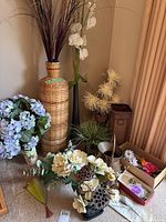 Wide shot of various artificial flower bunches and decorative woven stands grouped together on floor near wall and curtain, showing varied styles and sizes.