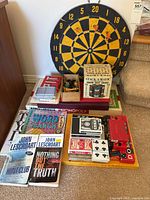 Photo showing dartboard, poker chip set, multiple decks of playing cards in various colors, and assorted board games and books stacked against a wall.