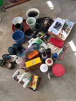 Overview of various garden supplies lying on garage floor including pots, sprays, shoes, gardening gloves, seed packets, and garden wire.