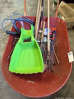 Red metal wheelbarrow containing various gardening tools and electric work lamp with blue extension cord.
