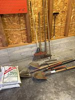 Wide view of shovels, rakes, pruner on floor with bag of fertilizer behind