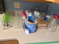 Photo showing various clear and green glass drinking glasses arranged on a countertop alongside glass bowls and a large clear glass jug with a red handle.