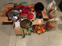 Wide shot showing open cardboard box containing various costume hats and headbands in red, black, gold colors with feathers and flowers; floral arrangements; and two glittery pumpkin decorations wrapped in plastic.