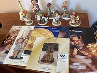 Photo of nine Hummel figurines, including various children in different poses and the Hummel decorative bell, arranged on a wooden surface with five Hummel booklets spread in the front.