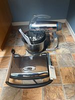 Sunbeam MixMaster stand mixer with stainless steel bowl containing beaters, a whisk and other attachments next to a closed Breville sandwich grill on tiled floor.