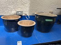 Three ceramic plant pots shown side-by-side on a blue surface outdoors, varying sizes, dark glossy finish with unglazed interior.