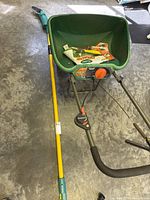 Green and orange fertilizer/grass spreader with large hopper, control dial, metal frame, and black handle grip.