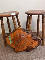 Two wooden stools with round tops and four turned legs shown behind a large wooden wall clock with bark edges and Roman numerals.
