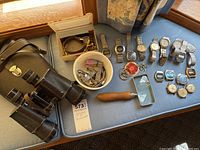 Photo showing binoculars, magnifying glass, assorted wristwatches laid out on a light blue surface, some watch bands in container, and keyrings.