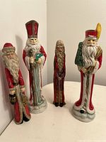 Front view of four vintage Santa figurines arranged side by side on a white surface showing details of faces, attire, and accessories.