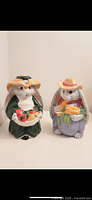 Front view of two lady bunny ceramic figurines, showing detailed clothes and accessories, one holding vegetables.