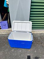 Blue plastic cooler with white lid, shown open revealing empty interior. Cooler is positioned on concrete near green storage unit door.