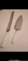 Photo of two cutting utensils: a serrated knife and a cake server, both with white resin handles and marked stainless steel blades, placed side by side on a flat surface.
