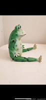 Side view of green frog figurine, showing seated posture, painted eyes and texture details.