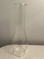 Tall clear glass hurricane lamp shade with narrow midsection and flared top edge shown on flat surface against plain background.