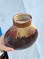 Oval-shaped vase held by hand, showing side angle with textured dark brown glaze and drip glaze finish