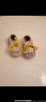 Front view of the pair of mini moccasins, showing the yellow lace bows and light cream color.