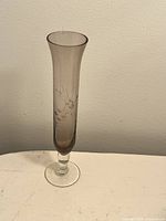 Clear photo of a tall, narrow small bud vase made of smokey glass with an etched design of a dragonfly and flower. Vase has a clear glass stem base.