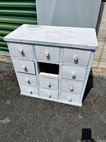 Front view of the small white wooden dresser with 11 drawers, one drawer missing in the middle row, showing wear and paint crackling.