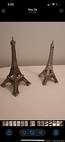 Photo showing two vintage brass Eiffel Tower figurines side by side on white surface against plain wall background.