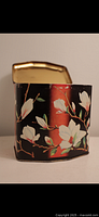 Front view of a vintage hinged metal tin with floral white magnolia flowers on red and black background, lid slightly open showing gold color inside.
