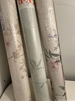 Three wallpaper rolls showing different floral and leaf patterns in neutral and pastel tones, leaning against a white wall.