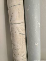 Close-up of two wallpaper rolls, one beige with abstract patterns, the other blue-gray with white birds