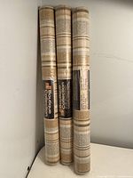 Three vertically standing unopened wallpaper rolls with beige striped pattern in plastic packaging.