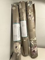Three rolls of wallpaper, two unopened, one opened, leaning against a white wall. Beige background floral pattern.