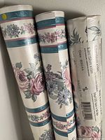Three wallpaper rolls standing upright, two unopened showing floral designs and one with leaf pattern label visible.