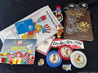 Overview of 1984 Olympics memorabilia laid out showing books, plates, pins, mascot figure, drinkware and fabric items