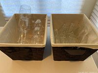Two brown woven rectangular baskets with beige cloth lining side by side, each filled with glasses. Left basket with 12 stemless glass champagne flutes, right basket with 8 stemless plastic wine glasses of two sizes.