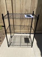 Front view of the assembled 3-tier black metal wire shelving unit placed outdoors on a concrete surface next to a wooden fence.