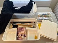 Black artist apron laid in a plastic bin with a white paint palette tray containing various brushes, a Handy Palette, and a small sponge.
